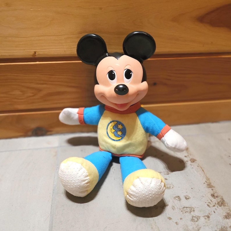 May include: A Mickey Mouse plush toy wearing a yellow and blue pajama set with a moon and stars graphic. The toy has black ears, a red collar, and white hands. The plush is sitting on a wooden surface.