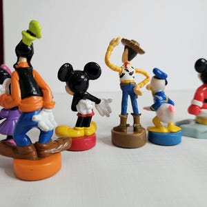 Op de afbeelding: Een set van zeven kleine plastic figuurtjes van populaire cartoonfiguren. De figuren omvatten Mickey Mouse, Goofy, Donald Duck, Woody uit Toy Story en drie andere figuren. De figuren staan allemaal op kleine voetstukken.