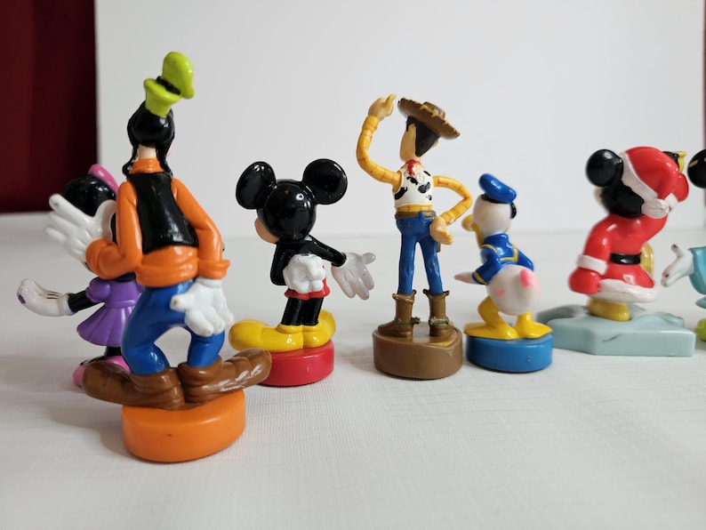 Op de afbeelding: Een set van zeven kleine plastic figuurtjes van populaire cartoonfiguren. De figuren omvatten Mickey Mouse, Goofy, Donald Duck, Woody uit Toy Story en drie andere figuren. De figuren staan allemaal op kleine voetstukken.