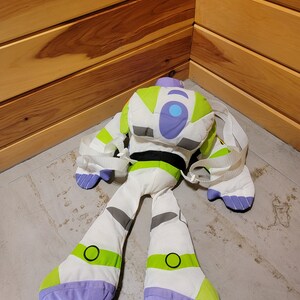 May include: A white and green plush toy of Buzz Lightyear from the Toy Story franchise. The toy is designed as a backpack with straps and has a purple and blue face with a large blue circle on the chest.