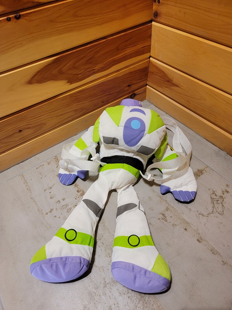 May include: A white and green plush toy of Buzz Lightyear from the Toy Story franchise. The toy is designed as a backpack with straps and has a purple and blue face with a large blue circle on the chest.