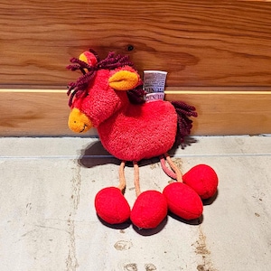 Peut inclure: Un jouet en peluche en forme de cheval rouge avec un bec jaune et des oreilles orange. Le jouet a des pieds rouges et une crinière et une queue rouge foncé. Une étiquette blanche est attachée au jouet.