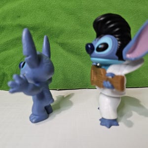 Peut inclure: Deux figurines bleues de Stitch. Un Stitch porte un costume blanc et a une coiffure en brosse noire. L'autre Stitch est debout, les bras tendus.