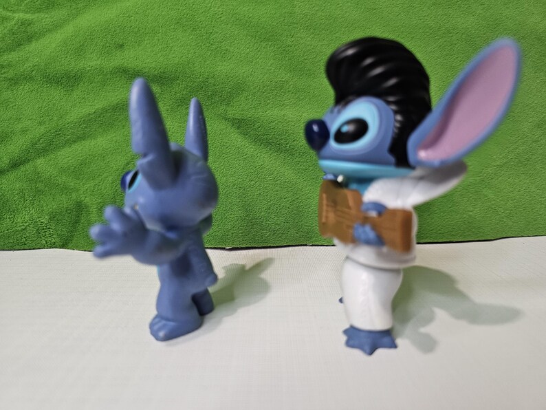 Peut inclure: Deux figurines bleues de Stitch. Un Stitch porte un costume blanc et a une coiffure en brosse noire. L'autre Stitch est debout, les bras tendus.