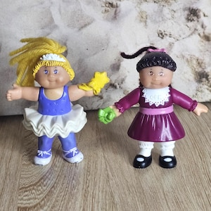 Peut inclure: Deux poupées vintage Cabbage Patch Kids. L'une porte un justaucorps bleu, un tutu blanc et des chaussures bleues, tenant une étoile jaune. L'autre porte une robe violette avec un col blanc et des chaussures noires, tenant un objet vert.