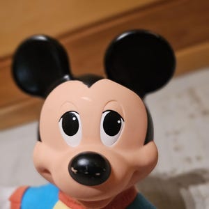 May include: A close-up of a Mickey Mouse doll. The doll has a black nose, black ears, and large, round eyes. It is wearing a blue and yellow shirt with red trim. The doll is a classic Disney character.