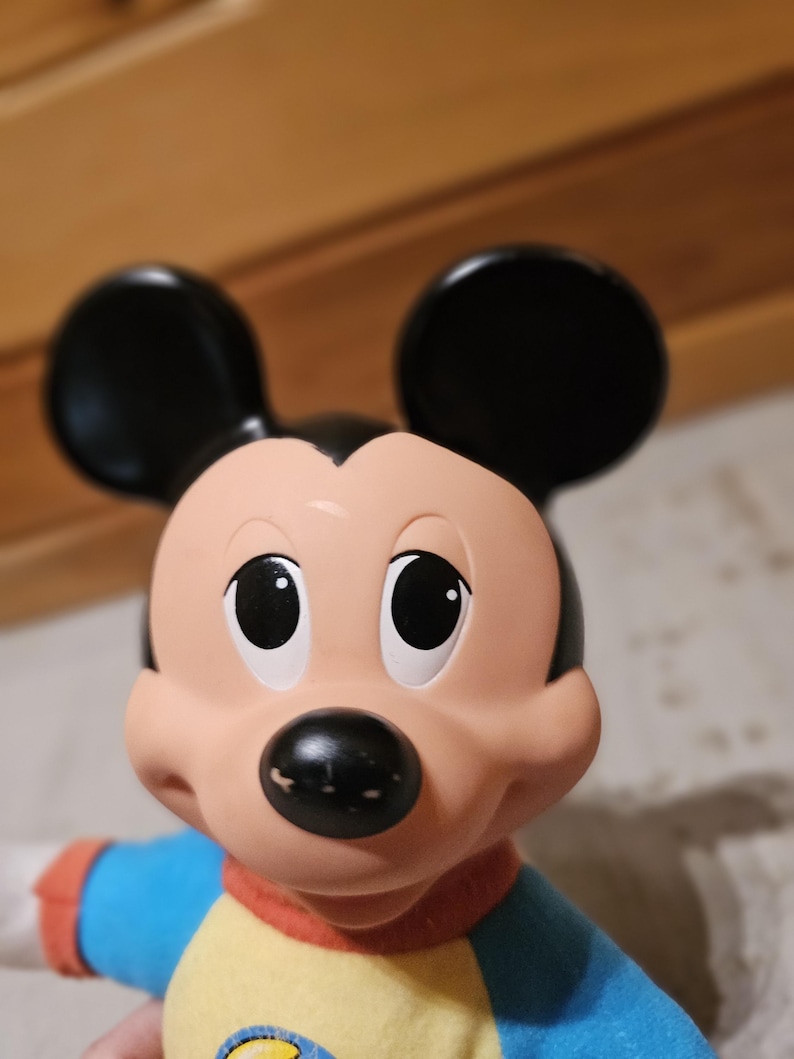 May include: A close-up of a Mickey Mouse doll. The doll has a black nose, black ears, and large, round eyes. It is wearing a blue and yellow shirt with red trim. The doll is a classic Disney character.