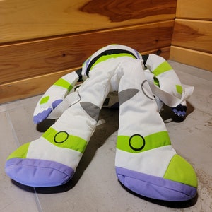 May include: A white, green, and purple plush toy shaped like Buzz Lightyear from the Toy Story movies. The toy has a backpack and is lying on its back with its legs in the air.