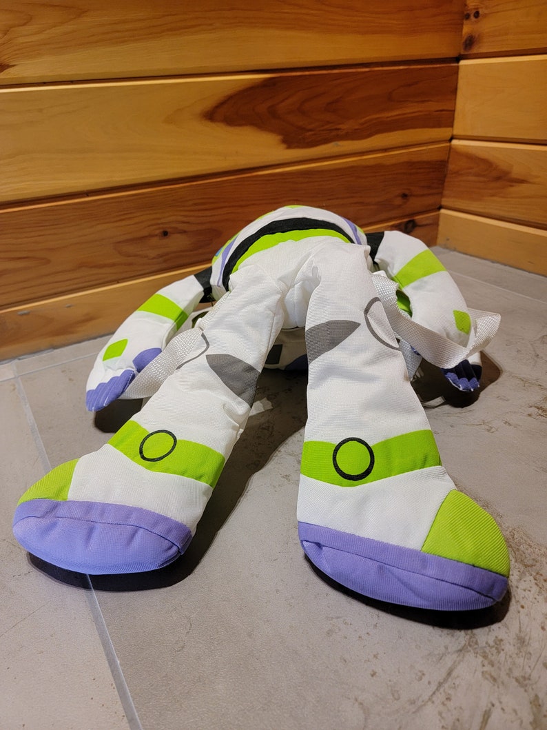 May include: A white, green, and purple plush toy shaped like Buzz Lightyear from the Toy Story movies. The toy has a backpack and is lying on its back with its legs in the air.