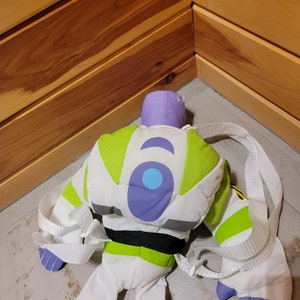 May include: A green, white, and purple plush toy backpack shaped like Buzz Lightyear from the Toy Story franchise. The backpack has a white strap and a black belt.
