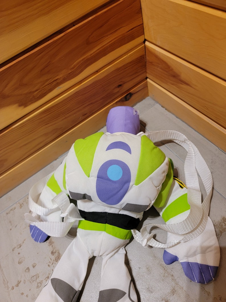 May include: A green, white, and purple plush toy backpack shaped like Buzz Lightyear from the Toy Story franchise. The backpack has a white strap and a black belt.