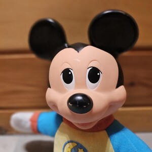 May include: A Mickey Mouse plush toy with black ears, a peach-colored face, and a black nose. The toy wears a yellow and blue pajama top with a moon and stars emblem. The sleeves are blue and red.