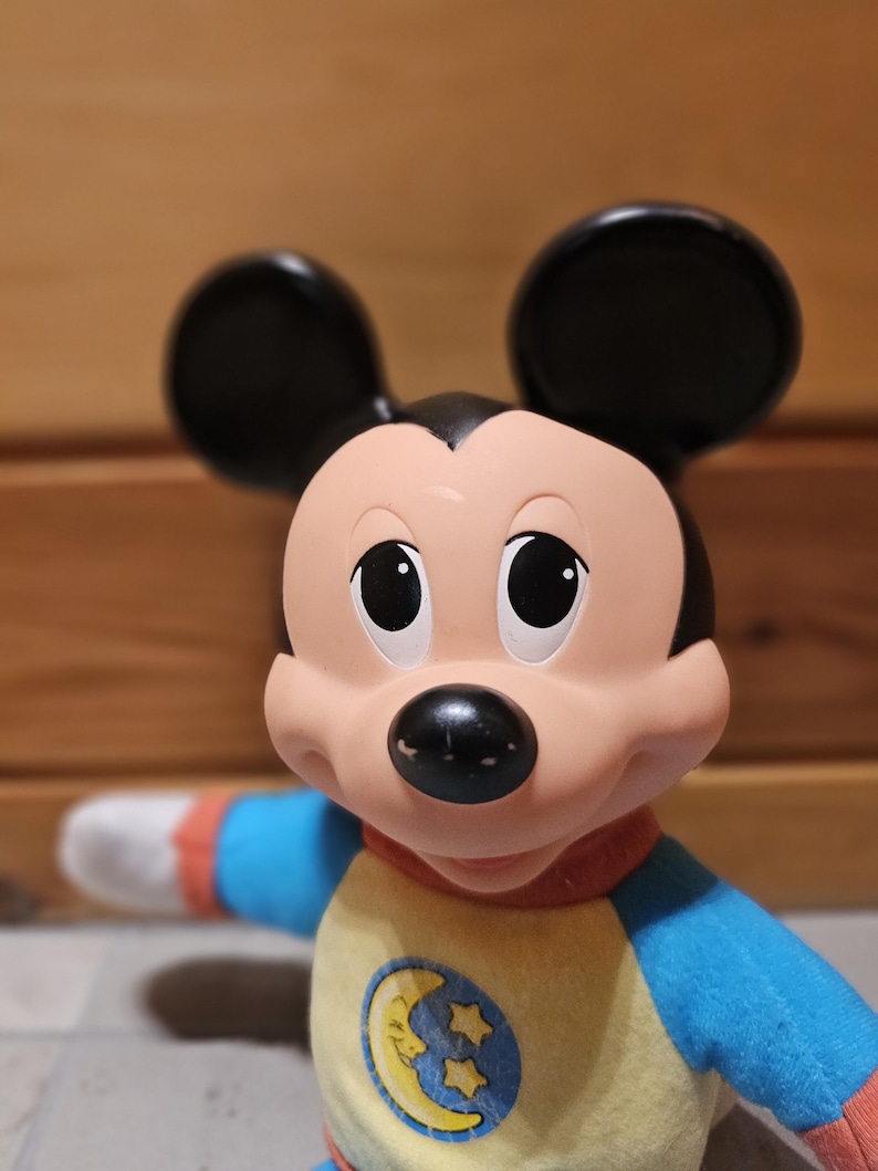 May include: A Mickey Mouse plush toy with black ears, a peach-colored face, and a black nose. The toy wears a yellow and blue pajama top with a moon and stars emblem. The sleeves are blue and red.