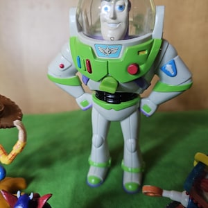 May include: A green and white Buzz Lightyear action figure with a clear plastic helmet and a purple face. The figure is standing with its arms crossed and has a red button on its chest.