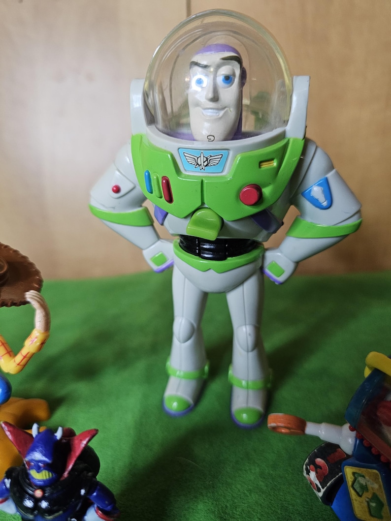 May include: A green and white Buzz Lightyear action figure with a clear plastic helmet and a purple face. The figure is standing with its arms crossed and has a red button on its chest.