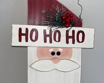 May include: A wooden Santa Claus face with a red hat, a white beard, and a red and white sign that says "HO HO HO".