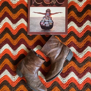 Könnte beinhalten: Ein Vintage-Vinyl-Schallplattenalbum mit dem Titel "Decade" mit einer Frau auf dem Cover und ein Paar braune Leder-Ankle-Boots. Die Artikel liegen auf einer farbenfrohen, gemusterten Decke in Orange, Braun und Creme.