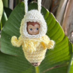 Può includere: Un costume da banana all'uncinetto con cappuccio bianco e corpo giallo. Una piccola bambola con i capelli castani e un viso sorridente indossa il costume.
