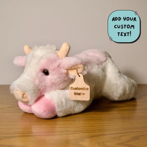 May include: A plush cow toy in white and pink. The cow has a light brown tag that says "Customize this!" and a blue rounded rectangle with the text "ADD YOUR CUSTOM TEXT!". The toy is lying on a wooden surface.