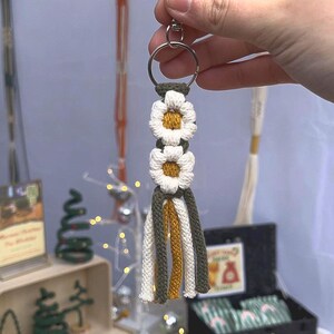 May include: A handmade macrame keyring featuring two white daisy flowers with yellow centres. The keyring has a silver ring and olive green, white, and gold tassels. Other macrame items are in the background.