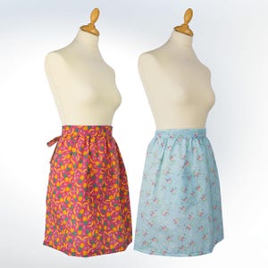 May include: Two aprons displayed on mannequins. One is a bright pink with a yellow and green fruit design. The other is light blue with a floral pattern and a pocket. Both are knee-length and have a gathered waist.