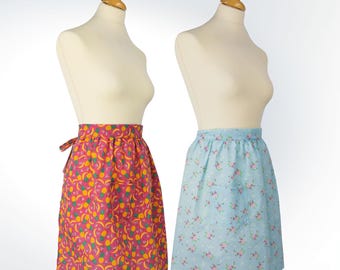 Puede incluir: Dos delantales en maniquíes. Uno es rosa vibrante con un estampado de frutas amarillas y verdes. El otro es azul claro con un estampado floral y un bolsillo. Ambos delantales son hasta la rodilla y tienen cintura fruncida.