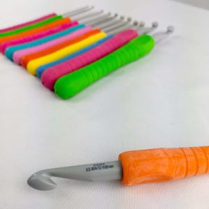 May include: A collection of colorful crochet hooks with ergonomic handles. The hooks are in various bright colors, including green, pink, yellow, and orange. One hook is in focus, showing the size: U.S. M/N-13/9.00 mm.
