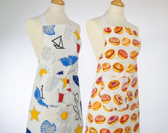 Puede incluir: Dos delantales en maniquíes. Uno es blanco con un diseño náutico que presenta un velero, un sol y conchas marinas. El otro delantal es blanco con un patrón de varios donuts. Ambos delantales tienen un bolsillo y correas blancas.