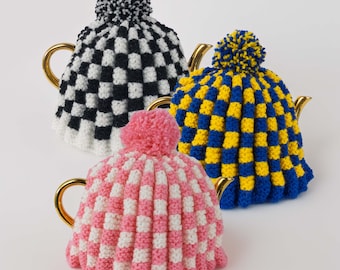 May include: Three tea cozies in a checkerboard pattern. One is black and white, another is blue and yellow, and the third is pink and white. Each cozy has a matching pom-pom on top and is placed on a gold-handled teapot.