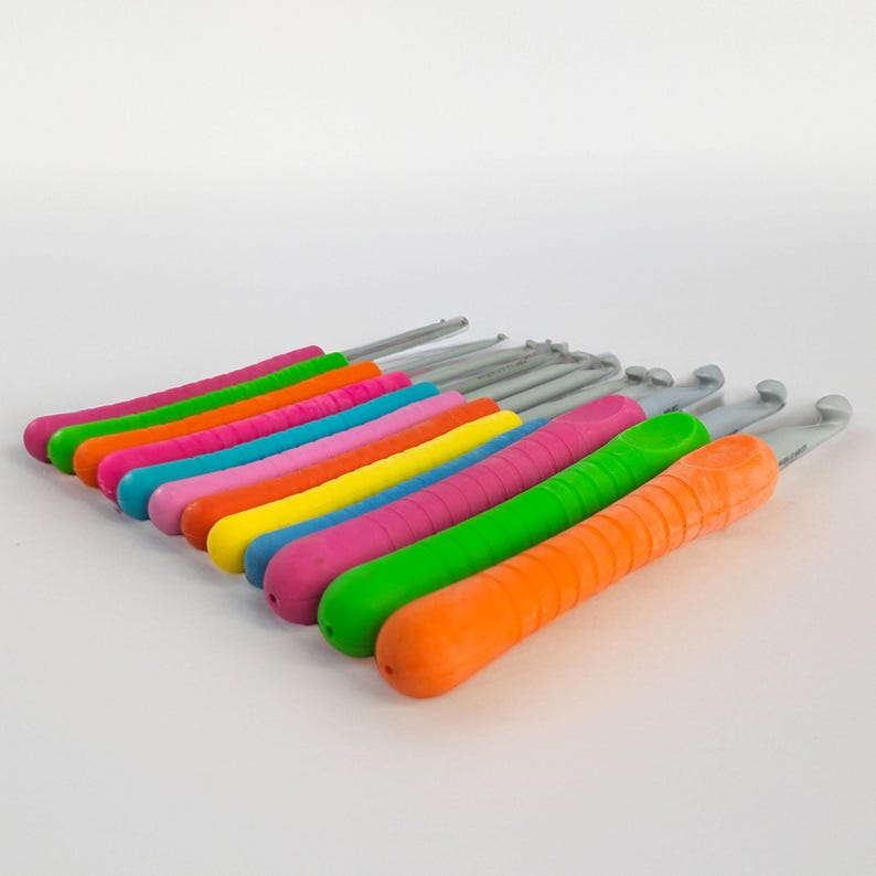 May include: A collection of crochet hooks with colorful, ergonomic handles. The handles are in a rainbow of colors, including pink, orange, green, yellow, and blue. The hooks are made of a silver-colored metal.