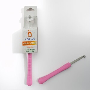 May include: Two pink crochet hooks with gray metal tips. One hook is still in its packaging, which includes the text "PONT 4.50 mm EASY GRIP". The other hook is lying separately.