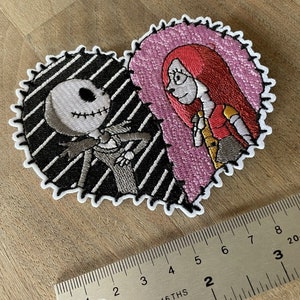May include: Embroidered patch featuring Jack Skellington and Sally from The Nightmare Before Christmas. The patch is shaped like a heart with a black and white striped background for Jack and a pink background for Sally.