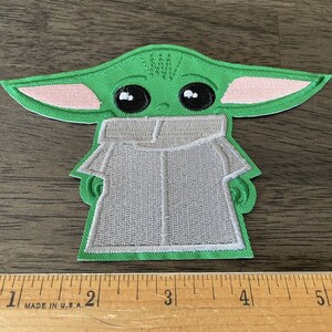 May include: Green and pink embroidered patch of a character from the Star Wars franchise, known as "The Child" or "Baby Yoda", wearing a grey hooded robe.