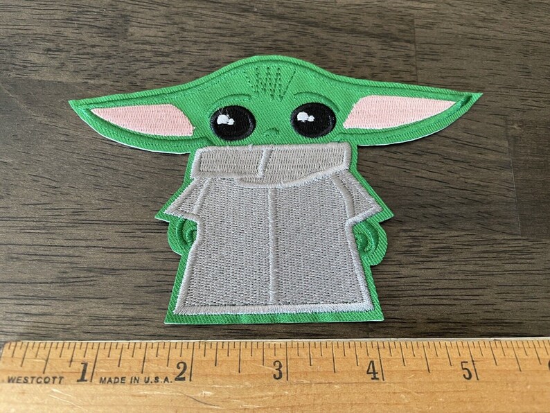 May include: Green and pink embroidered patch of a character from the Star Wars franchise, known as "The Child" or "Baby Yoda", wearing a grey hooded robe.