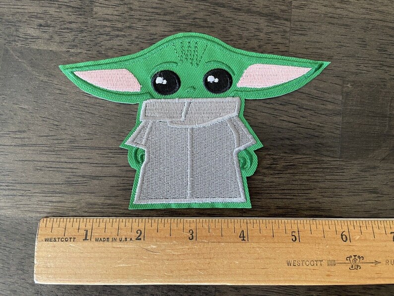 May include: Green and pink embroidered patch of a character from the Star Wars franchise, known as "The Child" or "Baby Yoda", wearing a grey hooded robe. The patch is approximately 2 inches tall.