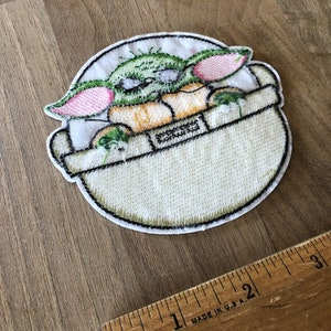 May include: Embroidered iron-on patch featuring a green, pink, and yellow cartoon character in a white pod. The character has large ears and is sitting in the pod.