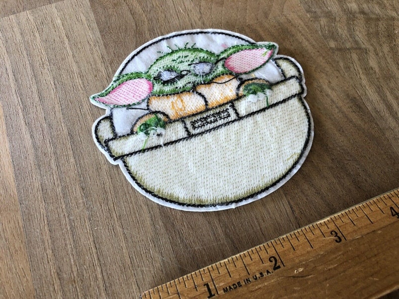 May include: Embroidered iron-on patch featuring a green, pink, and yellow cartoon character in a white pod. The character has large ears and is sitting in the pod.