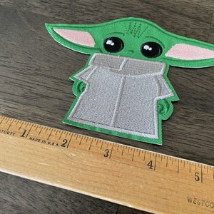 May include: Green and pink embroidered iron-on patch of a popular character from the Star Wars franchise. The character is wearing a gray hooded robe.