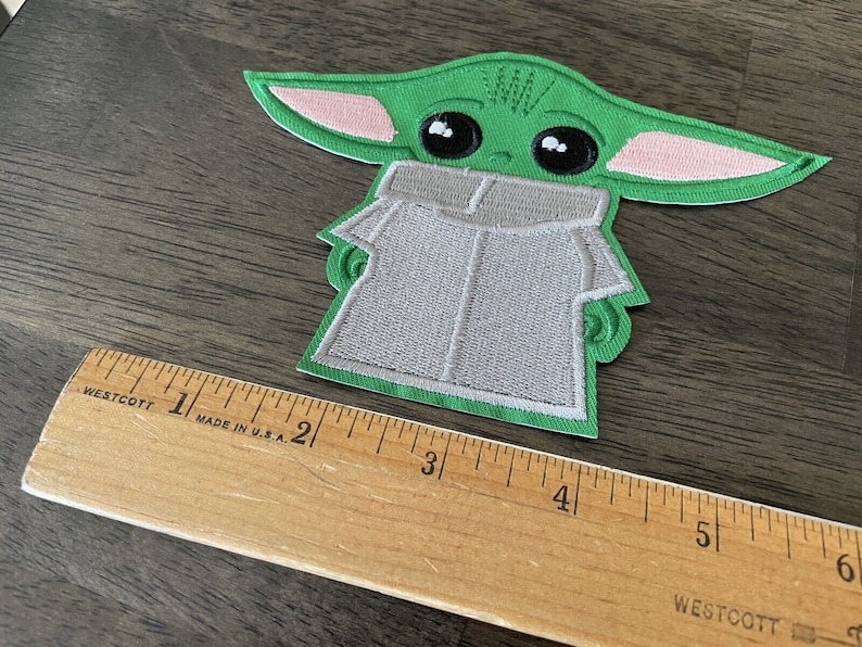 May include: Green and pink embroidered iron-on patch of a popular character from the Star Wars franchise. The character is wearing a gray hooded robe.