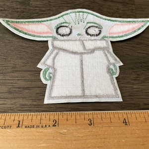 May include: A white embroidered patch of a green alien character with large eyes and pink ears. The character is wearing a white hooded robe. The patch is approximately 2 inches tall.