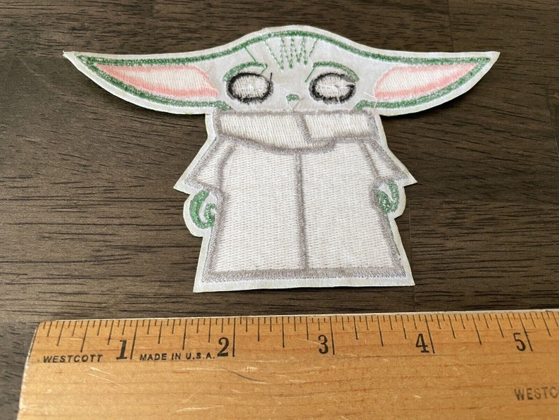 May include: A white embroidered patch of a green alien character with large eyes and pink ears. The character is wearing a white hooded robe. The patch is approximately 2 inches tall.