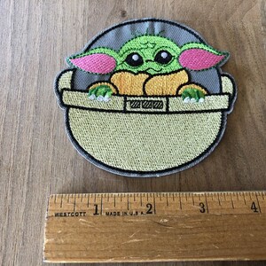 May include: Embroidered iron-on patch featuring a green Baby Yoda character from Star Wars, sitting in a floating pod. The patch is approximately 2 inches in diameter.