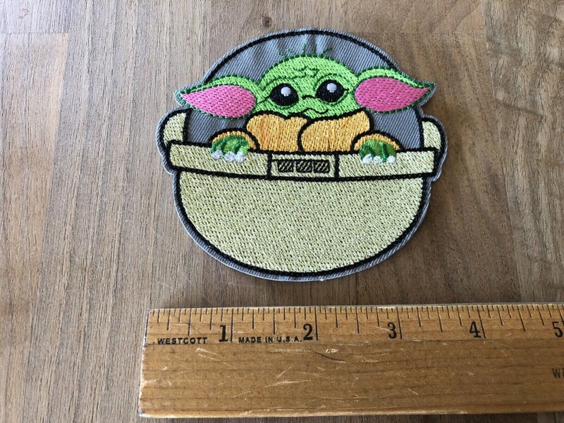 May include: Embroidered iron-on patch featuring a green Baby Yoda character from Star Wars, sitting in a floating pod. The patch is approximately 2 inches in diameter.