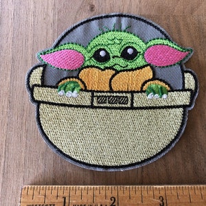 May include: Embroidered patch featuring a green character with large ears, pink ears, and big eyes, sitting in a gray and beige pod. The character is from the Star Wars franchise.