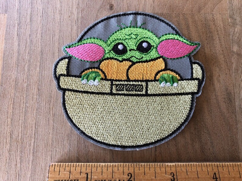 May include: Embroidered patch featuring a green character with large ears, pink ears, and big eyes, sitting in a gray and beige pod. The character is from the Star Wars franchise.
