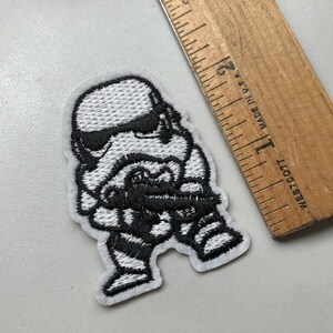 May include: A white and black embroidered patch of a Star Wars Stormtrooper character. The patch is approximately 2 inches tall.