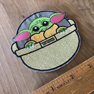 May include: Embroidered patch featuring a green, pink, and yellow character with large eyes, sitting in a gray and green pod. The character is from the Star Wars franchise.