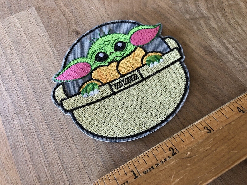May include: Embroidered patch featuring a green, pink, and yellow character with large eyes, sitting in a gray and green pod. The character is from the Star Wars franchise.
