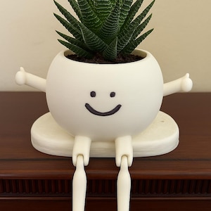 May include: A whimsical white planter with a smiling face, arms, and legs, holding a green succulent plant. The planter sits on a round base. A fun and unique decorative item for home or office.