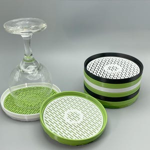 May include: A clear wine glass sits atop a green and white tray. Two additional trays, one green and white, and another stacked with black, white, and green bands, are also present. The trays have a slotted design.